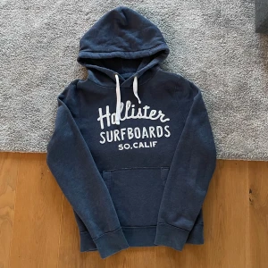 Mörkblå hoodie från Hollister - Srår XL men skulle hellre säga att den passar L. 184 som referens i bild. Mörkblå hoodie från Hollister med vit text 'Hollister Surfboards SO. CALIF' på bröstet. Klassisk känguruficka framtill, vita dragsnören i huvan och ribbade muddar. Perfekt för en avslappnad och cool stil.