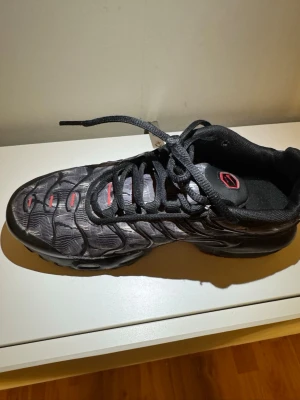 Nike Air Max Plus TN svart/röd manchester  - Nike Air Max Plus TN sneakers i svart och grått med röda detaljer. Skon har det klassiska vågmönstret på ovandelen, svarta skosnören och synlig dämpning i sulan. Materialet är en mix av mesh och syntet. TN-logga bak på hälen och ikonisk design.