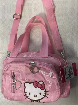 Hello Kitty Hand /  axelremsväska - Söt rosa axelremsväska med Hello Kitty-motiv från Sanrio. Väskan har två fack med dragkedja, justerbar axelrem och handtag. Dekorativa dragkedjor med Hello Kitty-formade hängen och mönster av små ikoner över hela väskan. Perfekt för dig som gillar kawaii-stil.
