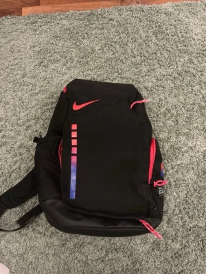 Nike Elite backpack ” kay yow” - Snygg svart ryggsäck från Nike med röda detaljer och dragkedjor. Framsidan har ett rött Nike-logotyp och färgade rutor i rött och blått. Ryggsäcken har flera fack och vadderade axelband, perfekt för skolan eller träningen.