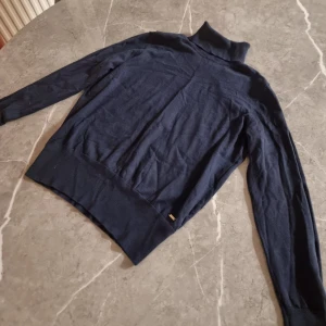 Mörkblå polotröja stickad - Mörkblå stickad polotröja med lång ärm och ribbade muddar. Tröjan har en hög krage och en liten metallbricka vid nederkanten. Perfekt för lager på lager och enkel att matcha med jeans eller kjol.