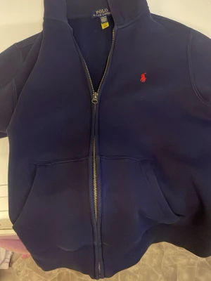 Marinblå halvzip från Polo Ralph Lauren - Snygg marinblå halvzip-tröja från Polo Ralph Lauren med röd broderad logga på bröstet. Tröjan har hög krage och dragkedja framtill. Perfekt för en clean och klassisk stil. Passar mig som är 158 men funkar för folk som är 160-165. Pris går att diskutera 