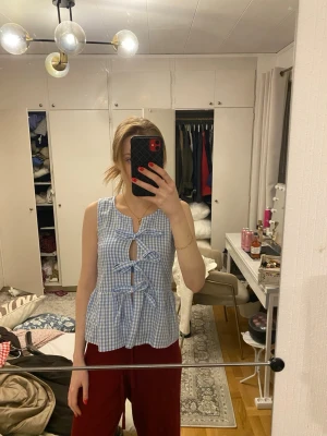 Blåvitrutig ärmlös topp med knyt - Supersöt ärmlös topp i blåvitt rutigt mönster med tre knyt framtill. Toppen har en lätt utsvängd passform och är perfekt för varma dagar. Tillverkad i ett tunt och luftigt bomullstyg som känns skönt mot huden.