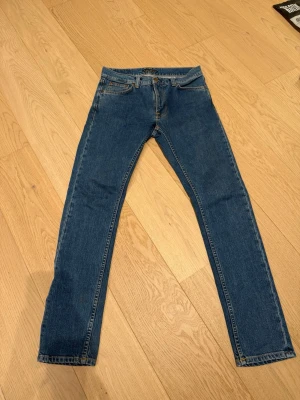 Blå raka jeans från Nudie Jeans - Klassiska blå jeans från Nudie Jeans med rak passform och fem fickor. Jeansen har normal midja, gylf med knapp och dragkedja samt kontrastsömmar. Tillverkade i slitstark denim, perfekta för en avslappnad och tidlös stil.