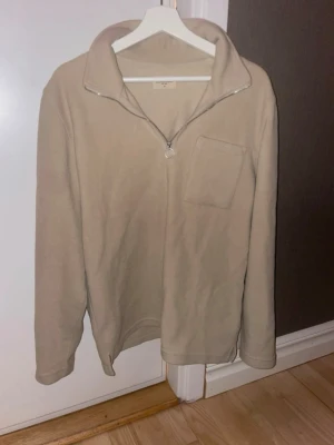 Beige fleece-tröja med halv dragkedja - Mysig beige fleece-tröja med halv dragkedja och hög krage. Tröjan har en bröstficka och långa ärmar. Perfekt för lager-på-lager och chill dagar. Enkel och stilren design som passar till det mesta.