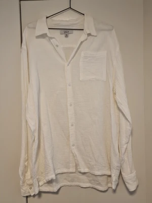 Vit långärmad skjorta från SMOG  - Klassisk vit skjorta från SMOG  i mjukt bomullstyg. Skjortan har lång ärm, bröstficka och knappar framtill. Perfekt oversized fit för en avslappnad look.