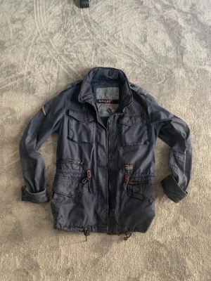 Mörkblå field jacket, size M - Snygg mörkblå field jacket med flera stora fickor framtill, axelklaffar och patch på ärmen. Jackan har dragkedja och detaljer i brunt samt en casual, normal passform. Perfekt för dig som gillar utility-stil och vill ha en jacka som sticker ut. Hör av dig vid minsta fundering och pris kan diskuteras! 🤩💯