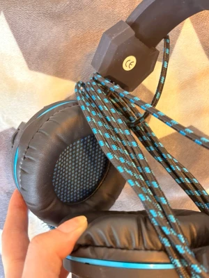 Svarta over-ear hörlurar med blå detaljer - Säljer ett par over-ear hörlurar med mjuka svarta kuddar och blå detaljer. Hörlurarna har ett flätat kabel med svart och blått mönster. Perfekt för gaming eller musiklyssning. Materialet är plast och konstläder.