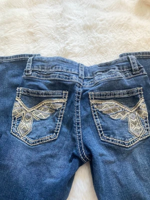 Blå bootcut jeans med broderade fickor - Snygga blå bootcut jeans med dekorativa broderier och detaljer i silver på bakfickorna. Jeansen har klassisk femficksdesign och kontrastsömmar. Perfekt för dig som gillar en unik och cool stil. Materialet är denim. Jeansen är insydda något för en tightare passform.💗