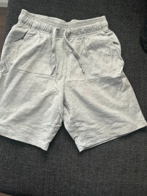 Grå mjuka shorts med snörning - Säljer ett par ljusgrå shorts med elastisk midja och snörning framtill. Shortsen har två sidofickor och är tillverkade i mjukt bomullsmaterial, perfekta för chill dagar eller träning. Klassisk och enkel design som passar till det mesta.