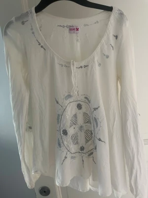 Vit boho-blus från Silke - Luftig vit blus från Silke med bohemisk känsla. Blusen har rund halsringning med knytband och broderade detaljer i blått och grått framtill samt på ärmarna. Tillverkad i ett tunt, lätt material som ger en avslappnad look.