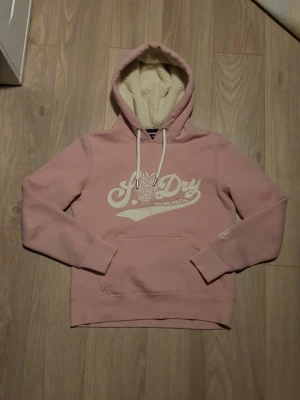 Rosa Superdry hoodie med teddyfoder - Mysig rosa hoodie från Superdry med vitt tryck på bröstet och stora snören i huvan. Insidan av huvan är fodrad med mjukt teddyfoder för extra komfort. Klassisk känguruficka framtill och ribbade muddar vid ärmar och nederkant.