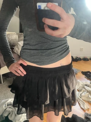 Svart volangkjol från KappAhl - Säljer en svart kortkjol från KappAhl med bred resår i midjan och dubbla lager volanger. Kjolen har en luftig och flowy känsla, perfekt för att styla med en enkel topp eller hoodie. Materialet känns lätt och bekvämt.