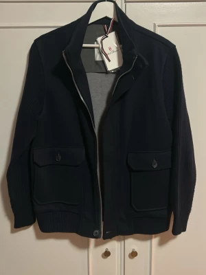 Mörkblå cardigan från Conte Of Florence - Snygg mörkblå jacka från Conte Of Florence med ribbade ärmar och nederkant. Jackan har två stora fickor framtill med lock och knapp, samt dragkedja hela vägen upp. Materialet är en mix av tyg och stickat, vilket ger en modern look.