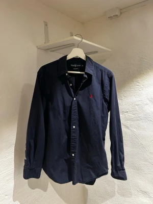 Mörkblå skjorta från Ralph Lauren - Klassisk mörkblå skjorta från Ralph Lauren med lång ärm och knappar framtill. Skjortan har den ikoniska röda logotypen broderad på bröstet och är tillverkad i mjuk bomull. Perfekt för en stilren och avslappnad look.