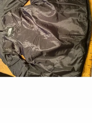 Svart väst från The North Face - Snygg svart väst från The North Face med klassisk design. Jackan är tillverkad i ett glansigt, lätt och vindtätt material. Perfekt för blåsiga dagar och enkel att packa ner i väskan. Diskret logga på bröstet och dragkedja framtill. 
