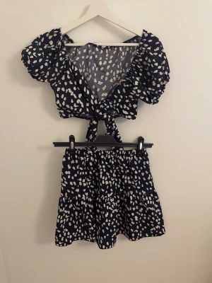 Mörkblå set med vita prickar - Säljer ett tvådelat set med mörkblå botten och vita prickar. Toppen har puffärmar och knyt framtill, medan kjolen är kort och har volanger. Setet är gjort i ett lätt och luftigt material som känns bekvämt mot huden. 