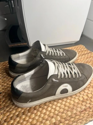 Grå sneakers Oliver Cabell  - Snygga grå sneakers från Oliver Cabell med vit sula och vita skosnören. Skorna har en vit detalj på sidan och svart hälparti. Tillverkade i mocka och läder för en clean och stilren look. Perfekta för dig som gillar minimalistisk design.