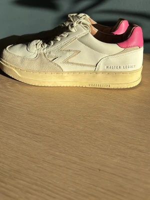 Knappt använda master legacy sneakers  - Snygga beige sneakers från Moaconcept med detaljer i mocka och läder. Skorna har en vit ovandel, beige sula och en cool rosa hälkappa. På sidan står det 'MASTER LEGACY'. Perfekta för dig som gillar stilrena och trendiga sneakers. Skorna är använda två gånger och ordinarie priset är ungefär 1900 kr. Skorna är storlek 39. 