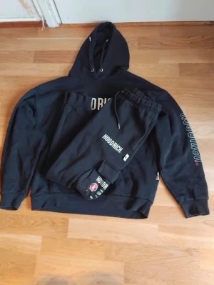 Svart Hoodrich set med tryck - Säljer ett svart Hoodrich set med hoodie och matchande byxor. Hoodien har stor vit logga på bröstet och tryck på ena ärmen. Byxorna har resår i midjan och tryck på benet. Båda delar har en avslappnad passform och är tillverkade i mjuk bomullsblandning.