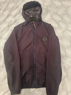 Mörklila vindjacka från Philipp Plein - Snygg svart vindjacka äkta från Philipp Plein med luva och dragkedja framtill. Jackan har ett diskret logomärke på bröstet och elastiska muddar vid ärmar och nederkant. Tillverkad i ett lätt och vattenavvisande material, perfekt för blåsiga dagar. Priset kan diskuteras samt mer bilder och ditaljer om jackan.