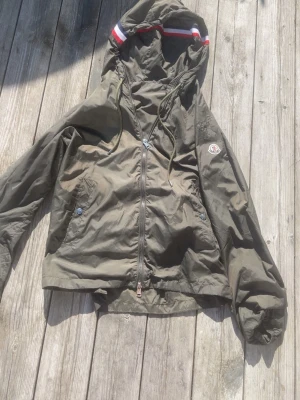 Olivgrön vindjacka från Moncler - Snygg olivgrön vindjacka från Moncler med huva och dragkedja framtill. Jackan har två fickor med tryckknappar och en ikonisk Moncler-logga på ärmen. Huva med dragsko och rödvit rand längs kanten. Perfekt för blåsiga dagar.