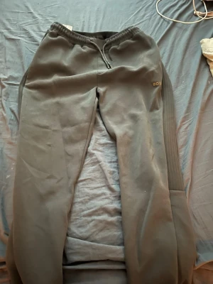 Grå mjukisbyxor från BOSS - Mörkgrå mjukisbyxor från BOSS med diskret logga på vänster sida. Byxorna har resår i midjan med dragsko och ribbade detaljer längs sidorna. Tillverkade i mjukt och bekvämt material, perfekta för chill dagar. Klassisk rak passform och stilren look.