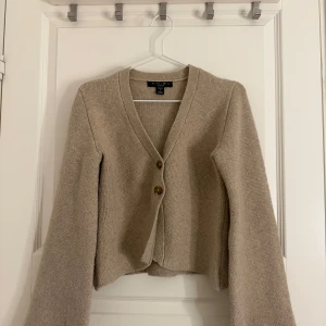 Beige kofta med knappar från New Yorker - Säljer en beige stickad kofta från Gina Tricot med två bruna knappar framtill. Koftan har v-ringning, lång ärm och en något croppad passform. Använts enstaka gånger. 