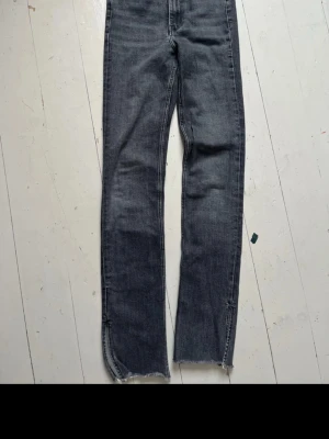 Grå raka jeans med slits nertill - Säljer ett par grå jeans med raka ben och slitsar nertill. Jeansen har råa, fransiga kanter vid bensluten för en trendig look. Materialet är klassisk denim i bomull och färgen är tvättad grå. Perfekta för en avslappnad och cool stil.