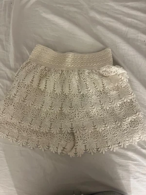 Vita virkade spets-shorts - Säljer ett par vita shorts med virkad spets och elastisk midja. Shortsen har ett detaljerat blommönster och vågig kant nertill, vilket ger en romantisk och bohemisk känsla. Perfekta för sommardagar eller festivalhäng.