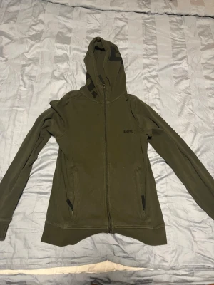 Olivgrön hoodie från Bench - Snygg olivgrön hoodie från Bench med dragkedja framtill och två fickor. Hoodien har en klassisk huva och Bench-loggan tryckt på bröstet. Perfekt för dig som gillar en enkel men stilren look. Materialet känns mjukt och bekvämt.