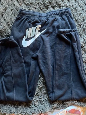 Svarta mjukisbyxor från Nike - Säljer ett par svarta mjukisbyxor från Nike med resår i midjan och muddar vid bensluten. Byxorna har stora vita Nike-loggor både fram och bak samt raka ben. Perfekta för chill eller träning, tillverkade i mjukt och bekvämt material.