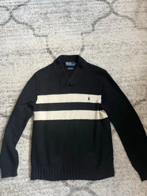 Svart stickad tröja Polo Ralph Lauren - Svart stickad tröja från Polo Ralph Lauren i 100% bomull. Tröjan har två breda vita ränder över bröstet och en liten broderad logga på vänster sida. Snygg krage med v-ringning och ribbade muddar vid ärmslut och nederkant.