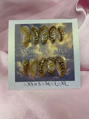 Opulent (S);Medium Almond - Handgjorda press-on naglar i storlek S: 15 mm, 11,5 mm, 12 mm, 11 mm, 9 mm . Naglarna har en nude bas med detaljerade guldfärgade mönster och små guldfärgade pärlor. Formen är medium mandel. Perfekt för dig som vill ha något unikt och glamoröst.