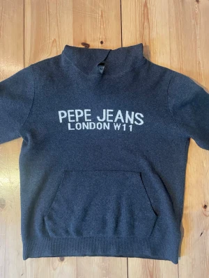 Pepe Jeans Stickad Hoodie - Mörkgrå stickad hoodie från Pepe Jeans med vit stilren text på bröstet tillverkad i mjukt material, perfekt för en avslappnad och cool stil. Storlek M Pris kan diskuteras!