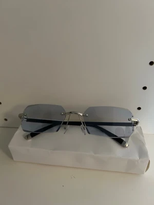 Silvriga solglasögon med blå ton - Snygga solglasögon med rektangulära, blåtonade glas och minimalistisk, silvrig metallbåge. Glasen är utan ram runt och har näskuddar för extra komfort. Perfekt accessoar för en clean och modern stil.