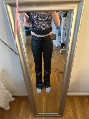Svarta raka byxor i skinnimitation - Säljer ett par svarta byxor i skinnimitation med rak passform och låg midja. Byxorna har klassisk design med knapp och dragkedja framtill. Perfekta för dig som vill ha en edgy look.