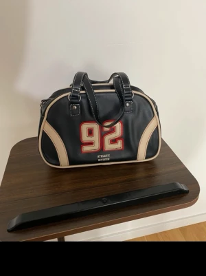 Svart handväska med beige detaljer - Cool svart handväska med beige kanter och handtag. På framsidan finns ett stort rött tryck med siffran 92 och texten 'Athletic Division'. Väskan har sportig look och är tillverkad i konstläder med vita sömmar. Perfekt för dig som gillar retrostil.
