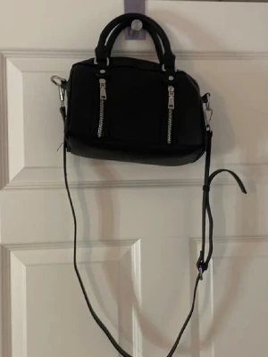 misako väska - BUDA!!!! Snygg svart handväska i boxig modell med två korta handtag och en lång justerbar axelrem. Väskan har två dekorativa silverfärgade dragkedjor framtill och stängs med dragkedja upptill. Perfekt för dig som gillar minimalistisk och stilren design.