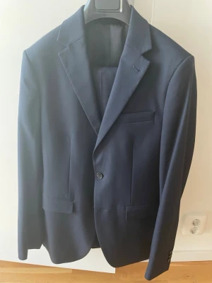 Mörkblå klassisk kavaj med byxor - Stilren mörkblå kostym  med två knappar och klassiska slag. Kavajen har tre fickor framtill och en slimmad passform som ger en modern look. Perfekt till skolavslutning eller student. Kostym är i slim fit, både kavaj och byxor, storlek kavaj 44/46. Byxor 46