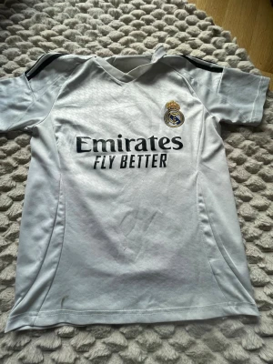 Real Madrid Mbappé matchtröja vit - Snygg vit Real Madrid matchtröja med Mbappé och nummer 9 på ryggen. Tröjan har klubbens emblem på bröstet och svart tryck med 'Emirates Fly Better' på framsidan. Korta ärmar och sportig passform, perfekt för fotbollsträning eller att bära till vardags.