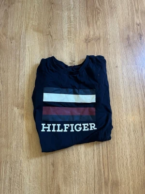 Svart Tommy Hilfiger t-shirt - Snygg svart t-shirt från Tommy Hilfiger med klassisk logga och färgblock i vitt, rött och marinblått på bröstet. Tillverkad i mjuk bomull och har en normal passform. Perfekt för en clean och stilren look.