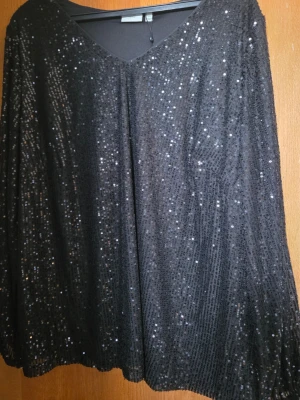 Svart glittrig paljettblus - Svart blus med v-ringning och långa ärmar, täckt av glittriga paljetter. Toppen har en loose passform och är gjord i ett syntetmaterial som ger ett snyggt fall. Perfekt för dig som vill ha något som verkligen sticker ut.