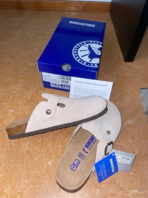 Beige mockatofflor från Birkenstock - Säljer ett par snygga beige tofflor från Birkenstock i mjuk mocka med klassisk rem och metallspänne. Fotbädden är ergonomisk och sulan är i kork och gummi för extra komfort. Perfekta för chill dagar hemma eller när du vill ha det bekvämt.