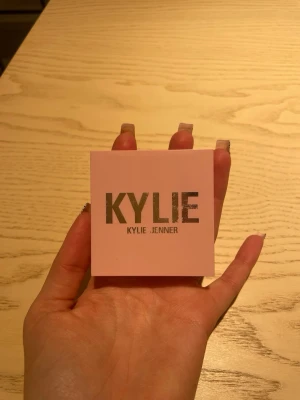 Kylie Jenner Pressed Blush Powder - Säljer ett pressat rouge från Kylie Jenner i färgen 'Winter Kissed'. Förpackningen är ljusrosa med silvrig text och kompakt fyrkantig form. Pudret ger en fräsch och naturlig look till kinderna och är lätt att applicera.