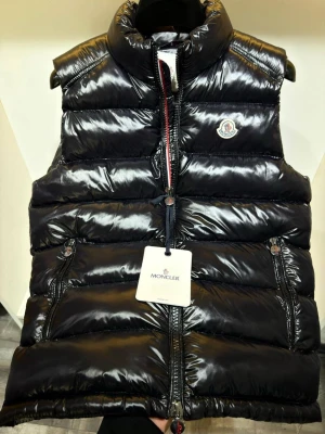 Moncler väst - Snygg svart dunväst från Moncler med glansig finish och hög krage. Västen har två dragkedjeförsedda fickor, Moncler-logga på bröstet och stängs med dragkedja framtill. Perfekt för lager på lager och streetwear-stil.