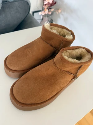 Uggs skor  - Mysiga bruna tofflor i mockaimitation med högre skaft och , beige foder i fuskpäls. Perfekta för att hålla fötterna varma hemma. Snygg söm längs sidan och stabil sula i gummi. Klassisk design som passar till chill hemma.