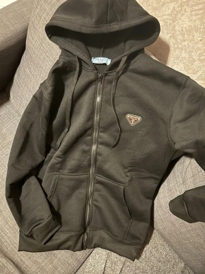 Svart hoodie med dragkedja från Prada - Svart hoodie från Prada med dragkedja framtill, huva med dragsko och klassisk logga på bröstet. Tillverkad i mjukt material och har två fickor framtill. Perfekt för en stilren och avslappnad look.