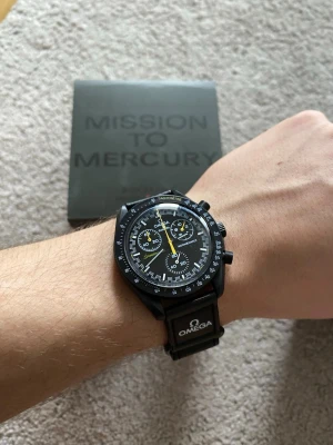 Omega x Swatch Mission to Mercury X Moon Remake - Snygg svart Omega x Swatch Mission to Mercury klocka med kronograf och detaljer i vitt och gult. Klockan har svart boett, svart armband med Omega-logga och tydlig tachymeterskala runt urtavlan. Levereras i originalbox.