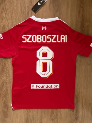 Liverpool Szoboszlai Fotbollströja - Säljer en röd Liverpool matchtröja från Adidas med den ungerska Szoboszlai på ryggen. Tröjan har vita detaljer och klubbmärke på bröstet. Tröjan har Champions League-märke på ärmen, sponsorlogga framtill och klassiska Adidas-ränder på axlarna. Tillverkad i funktionsmaterial, perfekt för fotboll eller träning. Tröjan är storlek S och är aldrig använd (lappen är fortfarande på).                          Skriv om ni har några funderingar!😄